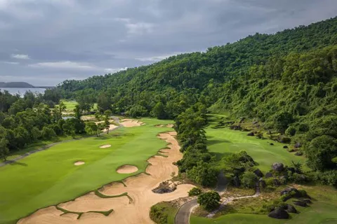 An aerial view of a green golf course with a lake and mountains in the background, showcasing Vietnam's diverse golfing lands