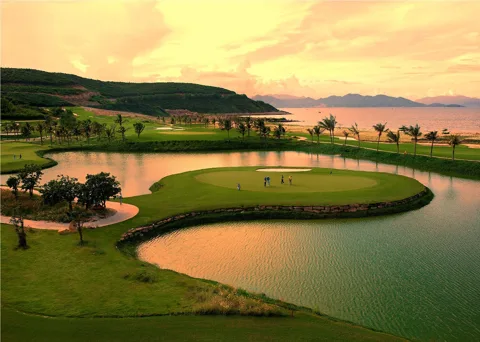 An aerial view of a green golf course with a lake and mountains in the background, showcasing Vietnam's diverse golfing lands